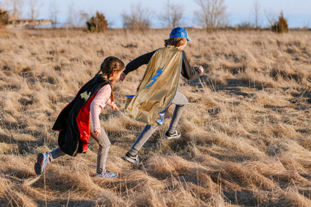 Two children playing in a field wearing Great Pretenders Superhero capes GP55273 and GP55785